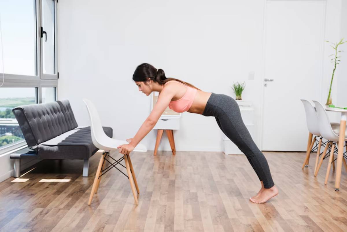 A woman in activewear performs an incline push-up using a chair, in a bright, modern living space with wooden flooring.