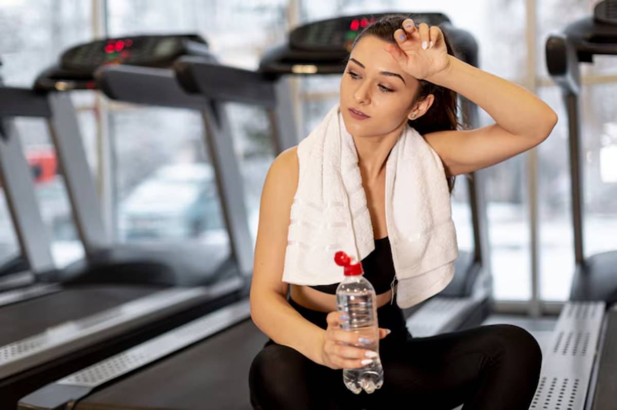 A woman in athletic wear sits on a treadmill, holding a water bottle and wiping her forehead with a towel, looking refreshed after exercise.
