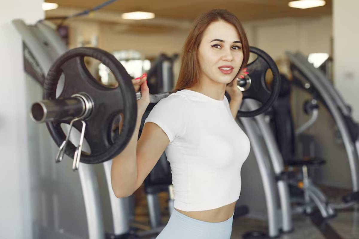 A young woman in a white crop top lifts a barbell as she exercises in a gym, focused on her workout with weight machines in the background.