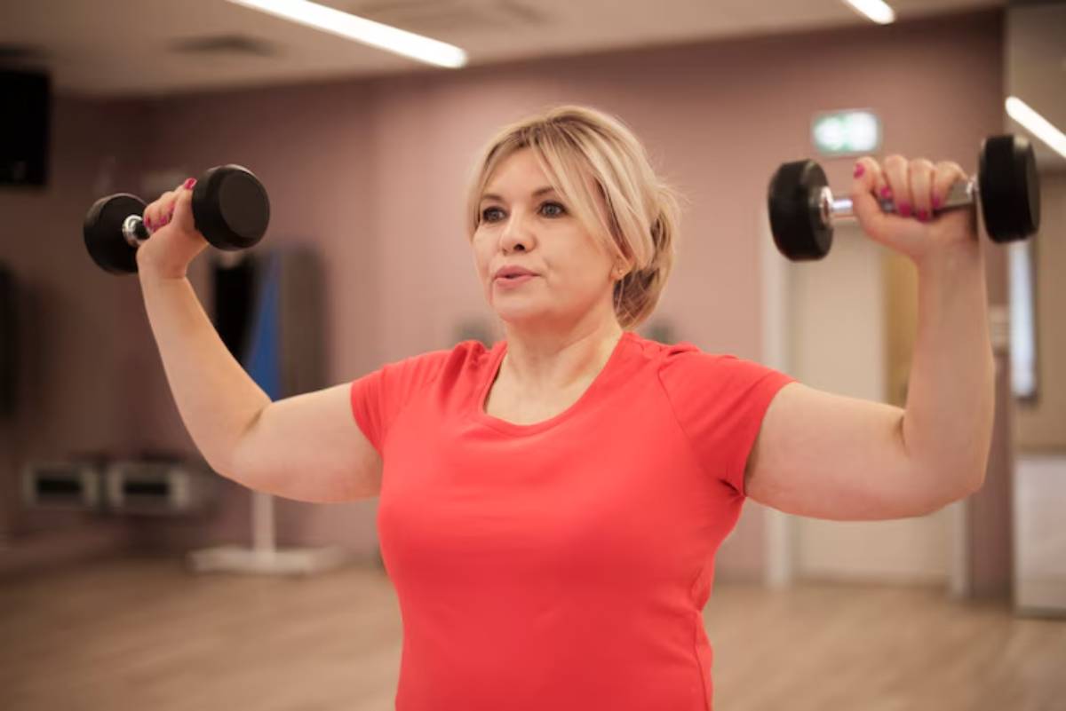 A woman in a red shirt lifts dumbbells above her head in a well-lit gym, demonstrating strength and fitness.