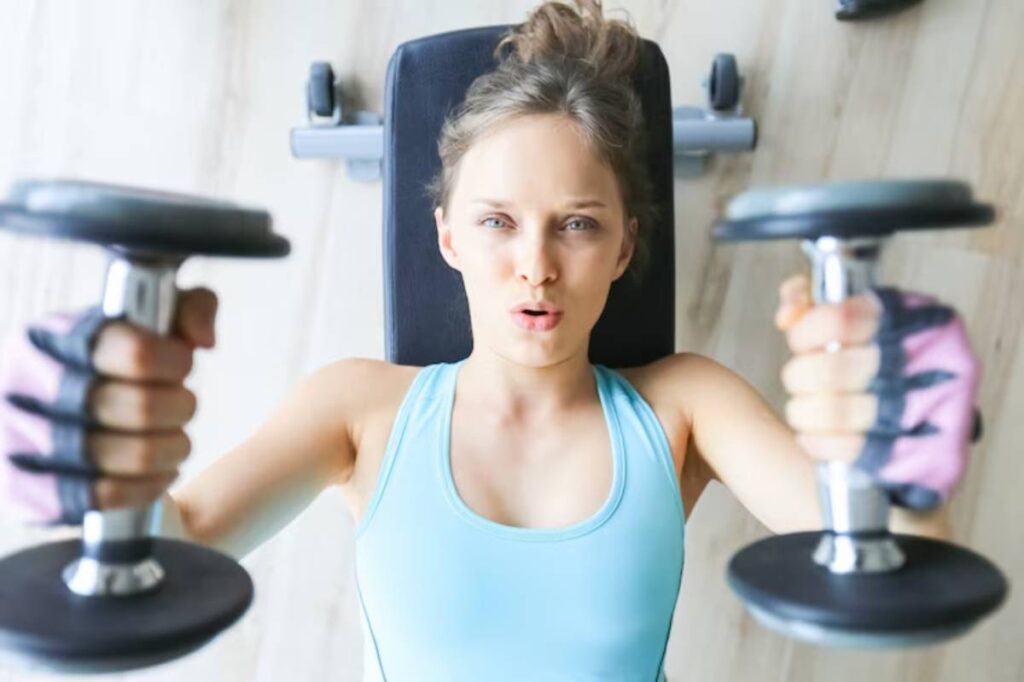 A woman is performing a weightlifting exercise on a bench, holding dumbbells in each hand, wearing a blue gym top and gloves.