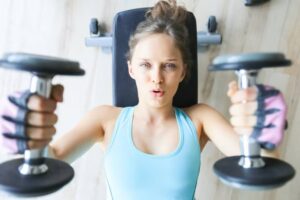 A woman is performing a weightlifting exercise on a bench, holding dumbbells in each hand, wearing a blue gym top and gloves.