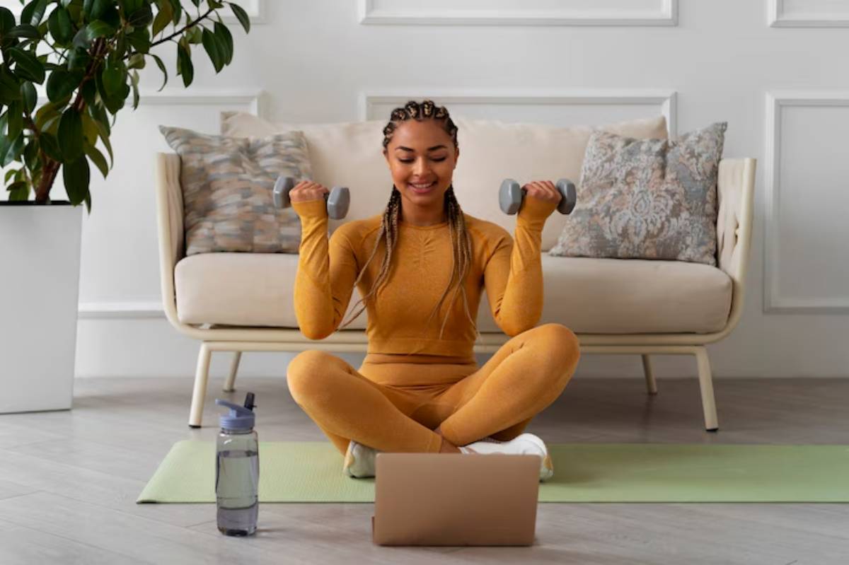  A person in an orange workout outfit lifts dumbbells while sitting on a yoga mat, with a laptop and a water bottle nearby.