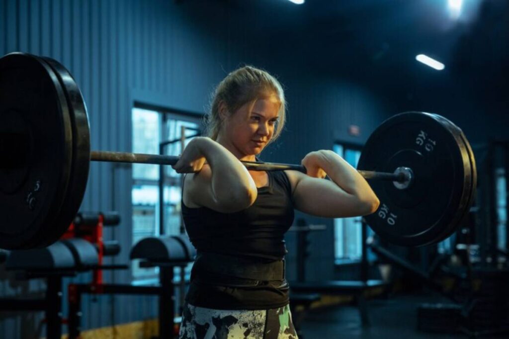 A person in workout attire performs a weightlifting exercise with a barbell in a dimly lit gym, showcasing concentration and strength.