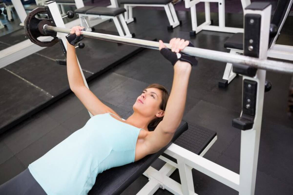 A person in a light blue tank top performs a bench press with a barbell in a gym setting, focusing on strength training.