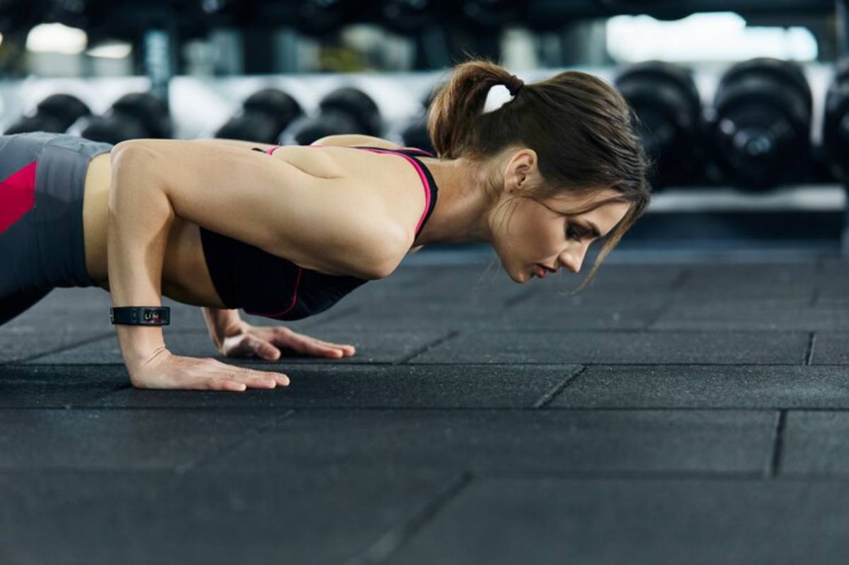 A woman in athletic gear performs a push-up on a gym floor, surrounded by weights in the background.
