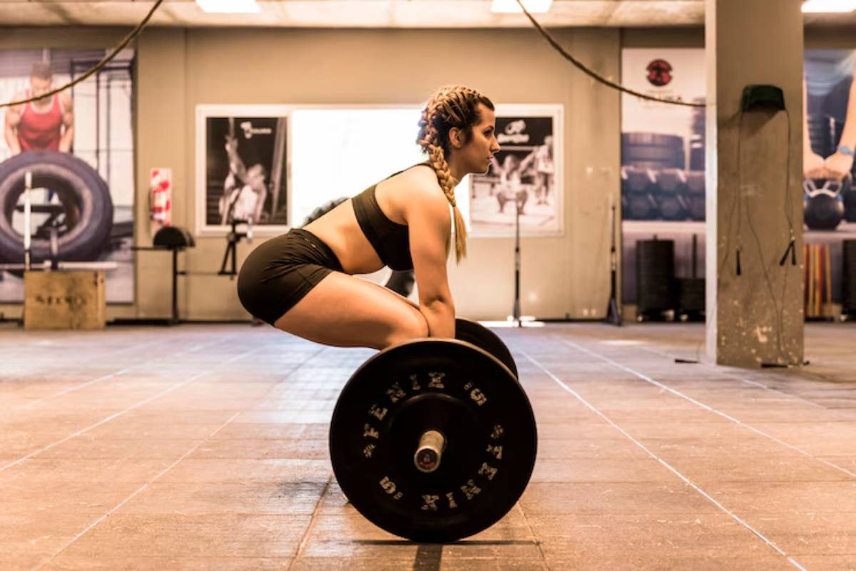 A person performs a deadlift in a gym, wearing a sports outfit, with weights on a barbell and gym equipment in the background.