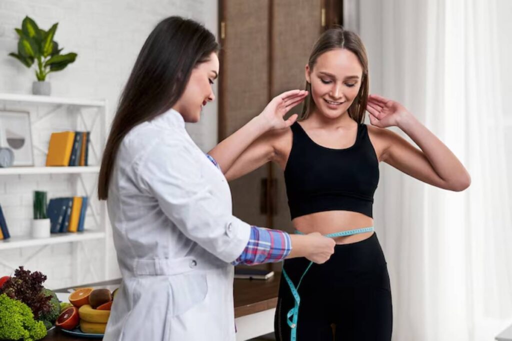 A healthcare professional measures the waist of a young woman in a fitness setting, surrounded by fresh fruits and vegetables.