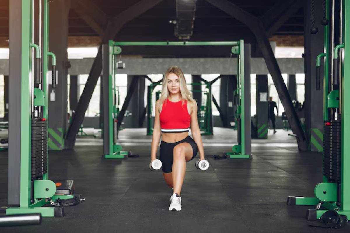 A person performs a lunge in a gym, holding dumbbells, showcasing strength training equipment and a spacious workout environment.