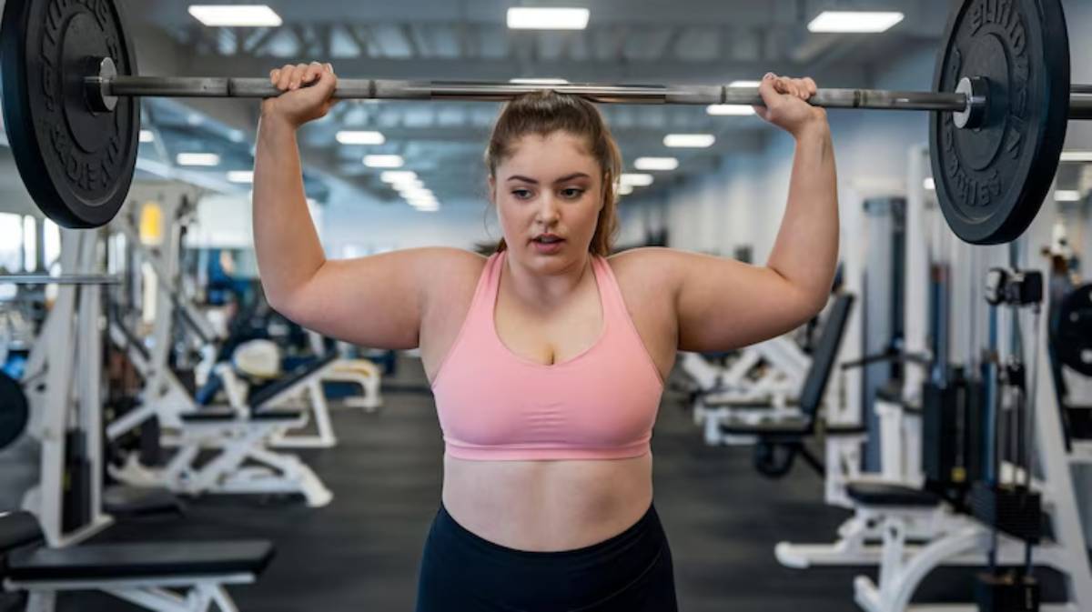 A woman in a pink sports bra lifts a barbell over her head in a bright gym, surrounded by fitness equipment.