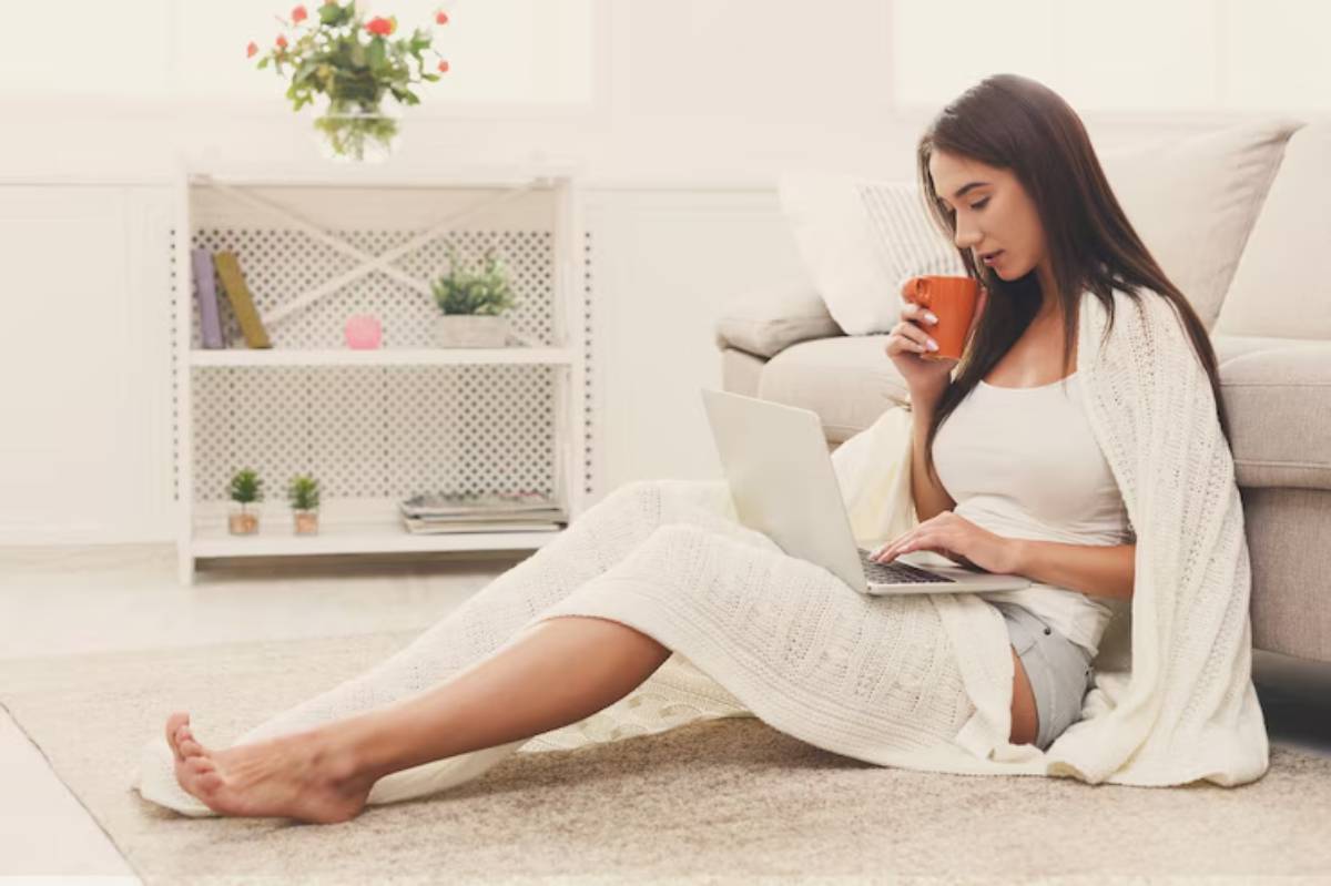 A woman wrapped in a cozy blanket sits on the floor with a laptop, sipping from an orange cup in a bright, airy room.